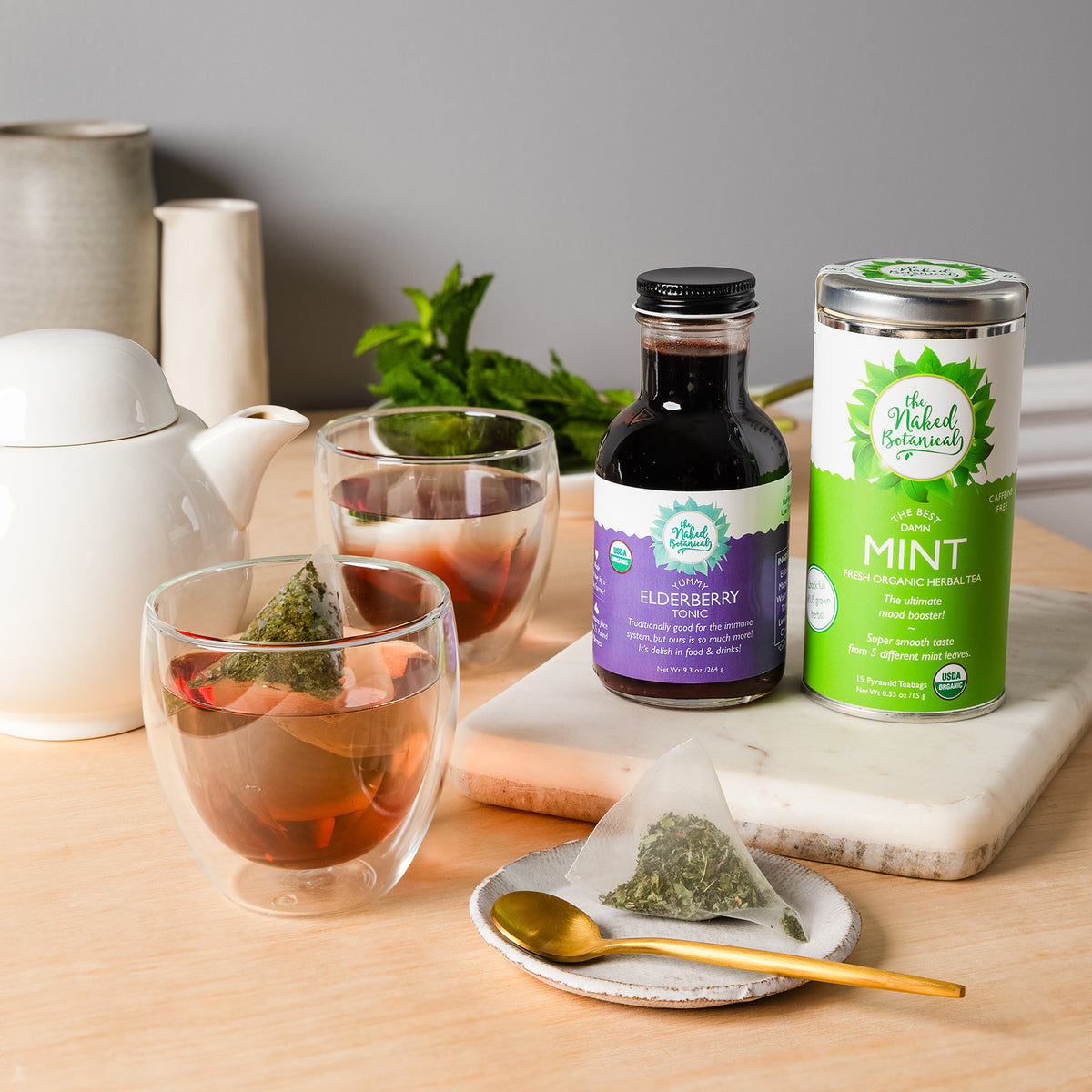 A styled tea setup on a light wood surface with a white teapot, two double-walled glass cups containing brewed tea, a small teabag in a plant based bag, and two bottles from The Natural Botanical :  an elderberry tonic bottle and a mint tea tin container   The cup contained brewed mint tea and a spoonful of elderberry tonic. The scene is set against a soft gray background with decorative ceramic vessels.